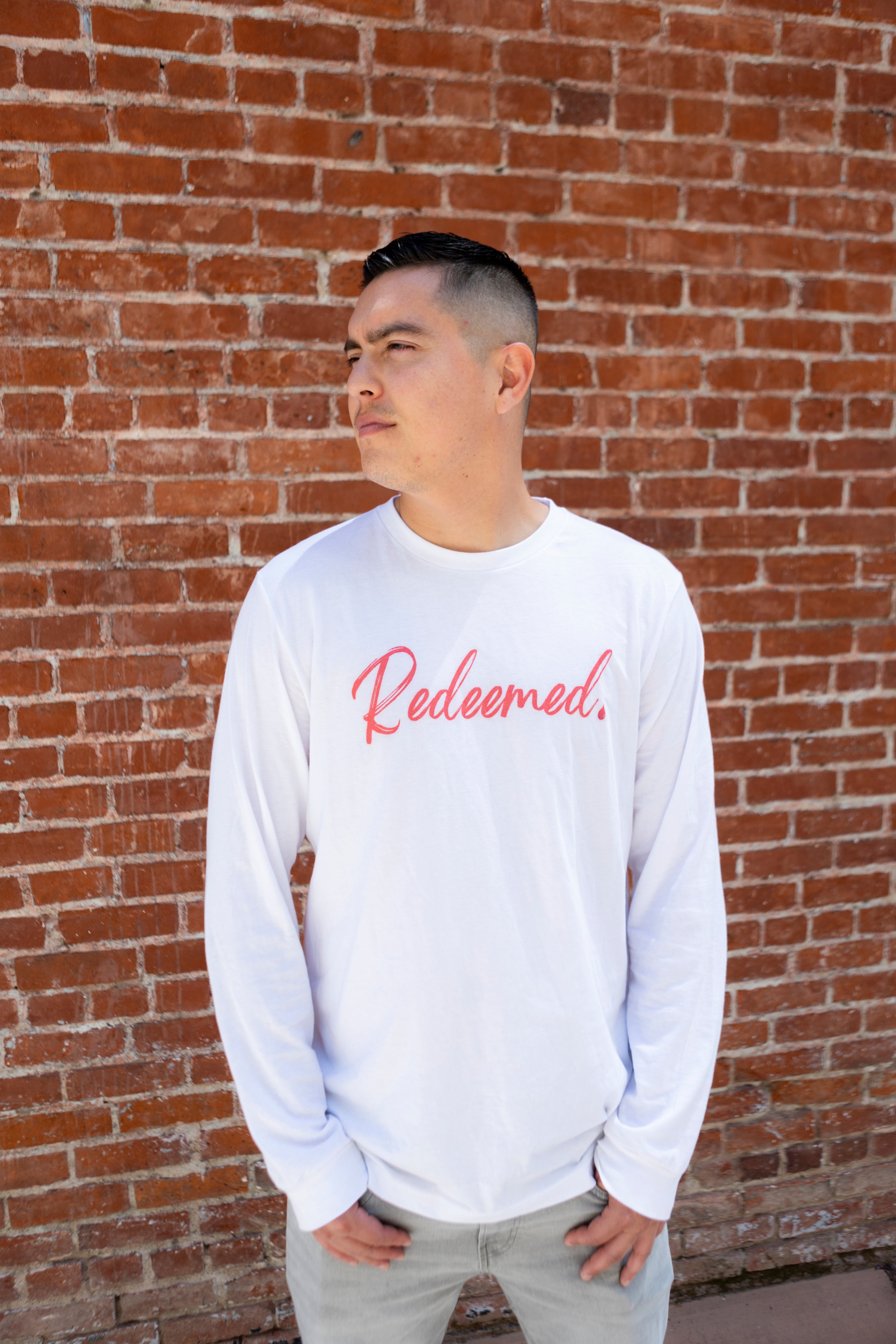 Person wearing a white long-sleeve shirt with 'Redeemed' text against a brick wall.